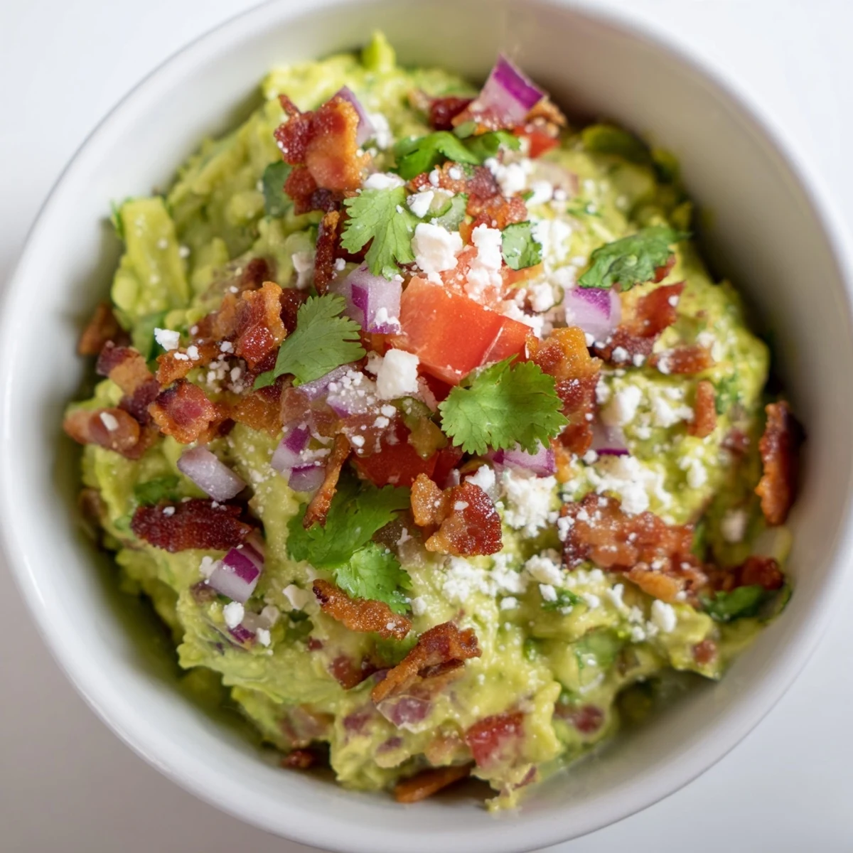 Creamy guacamole topped with crispy bacon and crumbled Cotija cheese, garnished with fresh cilantro and lime wedges for dipping.