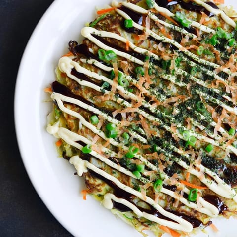 Steaming plate of delicious okonomiyaki pancakes, drizzled with sauce and sprinkled with bonito flakes.