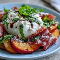 Creamy burrata, juicy peaches, and salty prosciutto on fresh greens with balsamic drizzle in summer salad.